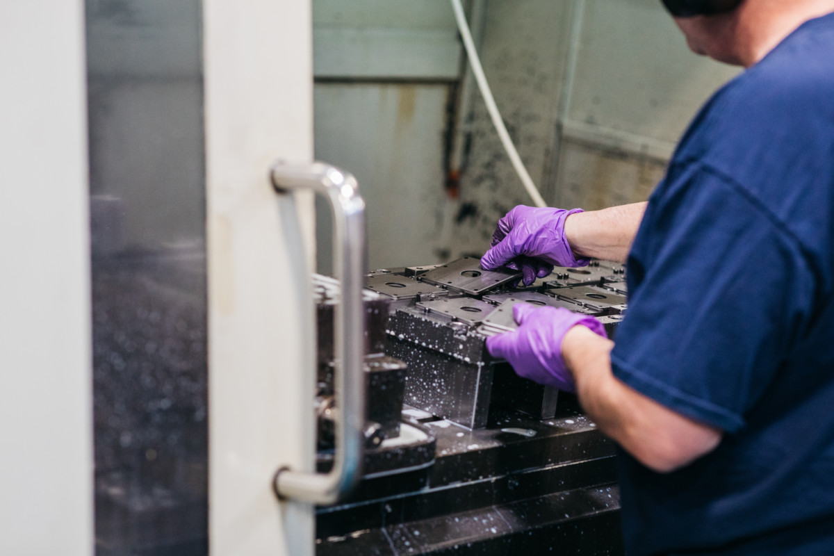 A person in a blue shirt and purple gloves removes stamped metal components from a machine.